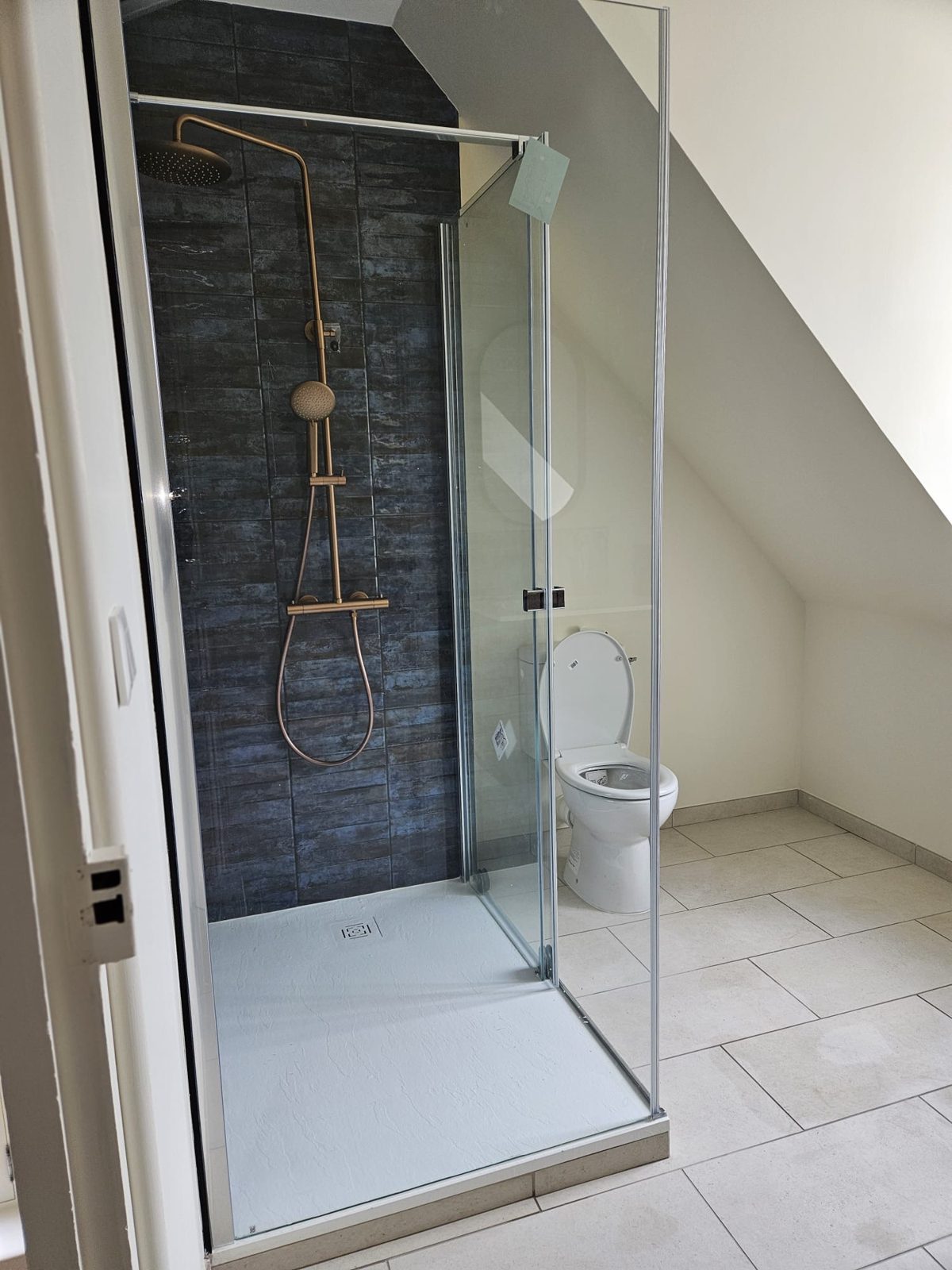 Douche avec paroi vitrée, receveur extra-plat blanc et colonne de douche cuivre dans une salle de bain sous combles