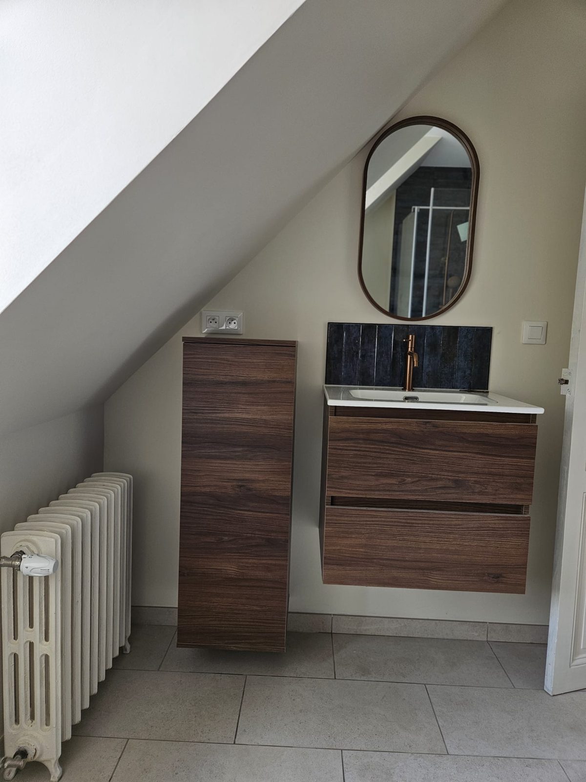 Meuble vasque en bois, robinet cuivre et miroir ovale dans une salle de bain rénovée sous combles