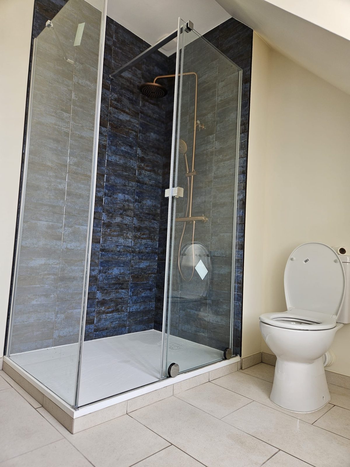 Salle de bain sous combles rénovée à Rennes : douche vitrée avec faïence bleue, WC et carrelage beige grand format